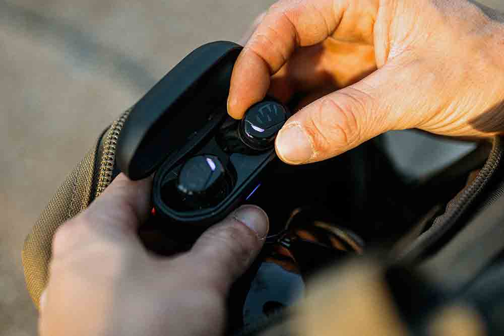 Hands opening a black charging case containing Walker's Suppressor Earbuds, showcasing the Bluetooth hearing protection device for hunters and shooters in an outdoor setting.